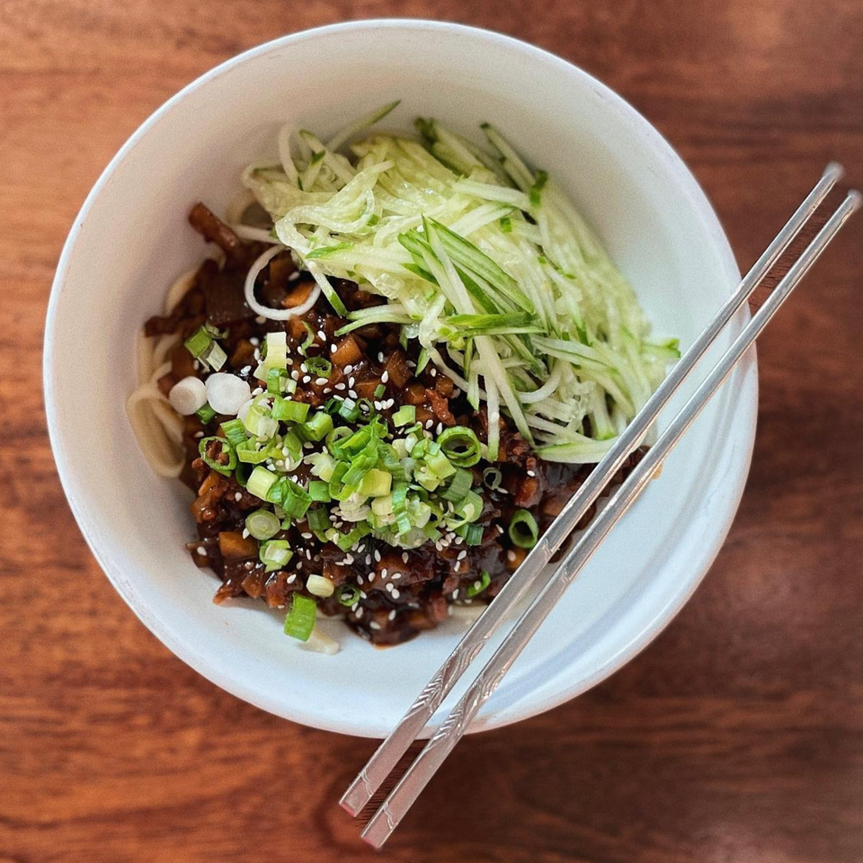 A bowl of Asian noodles with dark sauce, green onions, sesame seeds, and shredded cucumber, with chopsticks.