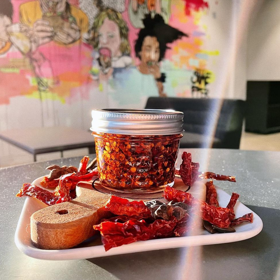 A jar of chili crisp and dried red chilies on a white plate, with a colorful mural in the background.