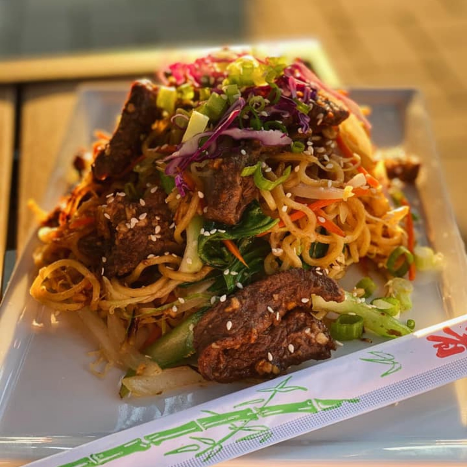 Asian noodles with beef, vegetables, sesame seeds, and chopsticks on a white plate.